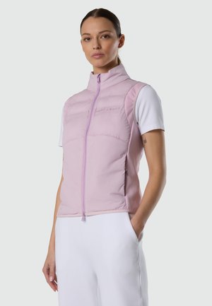 Frau trägt lavendelfarbenes, ärmelloses Steppwesten über weißem kurzärmligem Hemd, steht mit einer Hand in der Tasche vor einfachem Hintergrund.