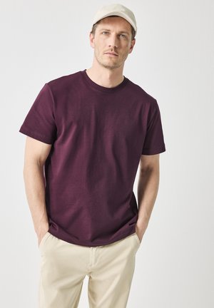 T-shirt a maniche corte bordeaux realizzato in cotone, con design a collo rotondo. Il modello indossa pantaloni beige e un cappellino di colore chiaro.