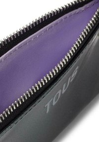 Cartera de cuero negro con un interior de tela púrpura, que cuenta con un cierre de cremallera plateado y el logo en relieve "TOUS" en el lado exterior.