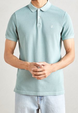 Mężczyzna w jasnozielonej koszulce polo z krótkim rękawem, kołnierzykiem i guzikami, z rękami złączonymi z przodu, w jasnoniebieskich dżinsach.