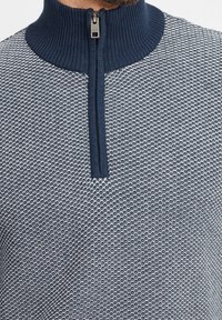 Pull en maille avec un col côtelé haut, doté d'une fermeture éclair et d'un motif texturé dans des tons marine et gris clair.