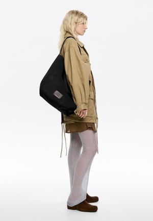 Borsa a rete nera con una patch di branding, indossata su una spalla. Il modello indossa una giacca khaki, una gonna marrone e collant in rete argentata.
