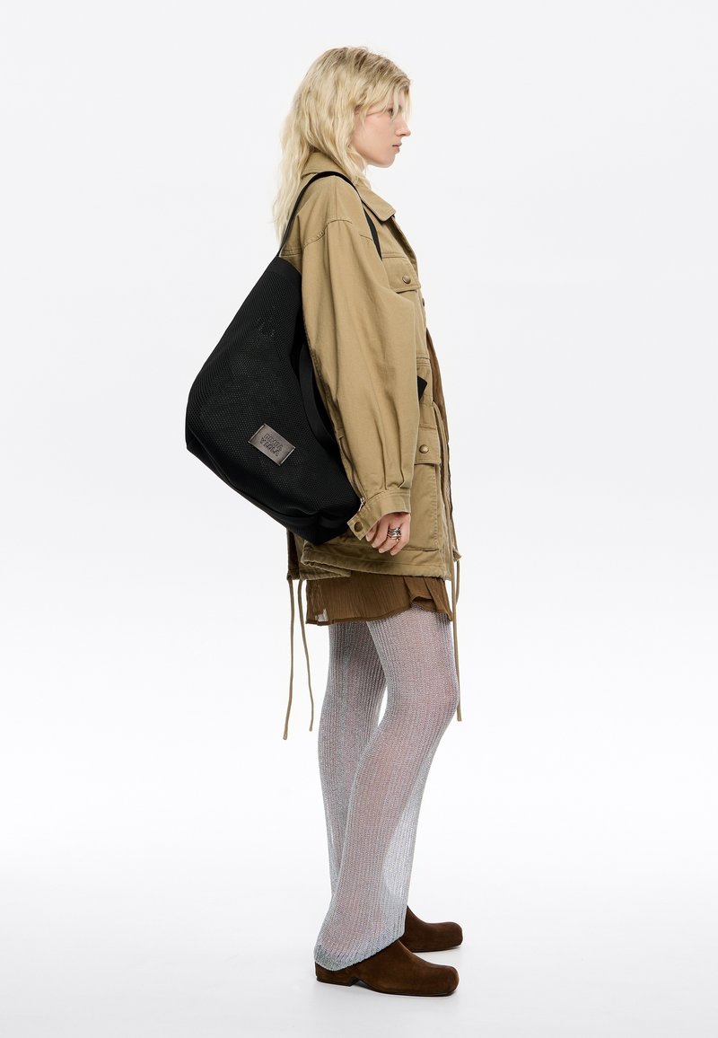 Zwarte mesh tote bag met een merkpatch, gedragen op één schouder. Het model draagt een kaki jas, een bruine rok en zilveren mesh panty’s.