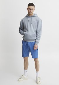 Grå hættetrøje med en frontlomme, parret med blå shorts. Modellen har hvide sneakers og hvide sokker på, hvilket viser afslappet atletisk tøj.