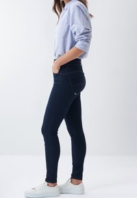 Ciemnoniebieskie rurki z tylnymi kieszeniami, zestawione z jasnoniebieską koszulą na guziki i białymi trampkami. Gładka tekstura denimu.