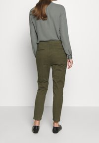 Femme debout, de dos, portant un pantalon vert olive, un haut gris à manches longues et des chaussures noires sans lacets sur un fond uni.