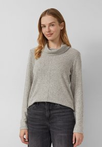 Frau mit hellbraunem Haar, die einen grau gemusterten Schalkragenpullover und schwarze Jeans trägt, steht vor einem schlichten, hellgrauen Hintergrund.