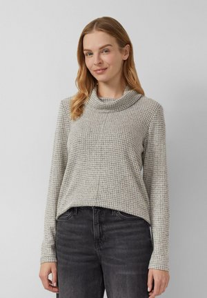 Femme aux cheveux châtains clairs portant un pull à col roulé gris à motifs et un jeans noir, se tenant devant un fond gris clair uni.