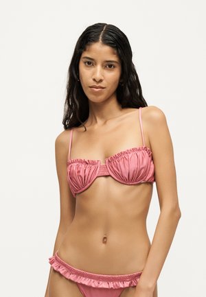 Femeie cu păr lung și închis la culoare poartă un costum de baie roz cu volane, stând în fața unui fundal deschis la culoare, privind către cameră.