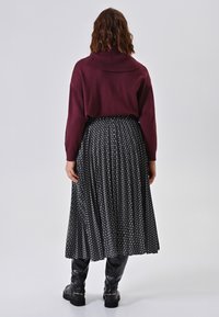 Bordeauxfarbener Strickpullover mit einem gerippten Kragen, kombiniert mit einem gefalteten Rock in Schwarz-Weiß-Muster und kniehohen schwarzen Stiefeln.