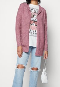 Gemaroonde cardigan met capuchon en een getextureerde afwerking, met twee borstzakken, gedragen over een graphic t-shirt en gecombineerd met versleten lichtblauwe jeans.