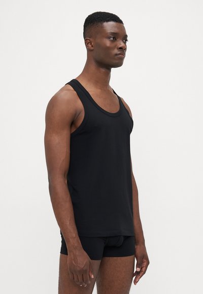 Mann in schwarzem Tank-Top und schwarzen Boxer-Shorts steht nach rechts gerichtet vor einem schlichten weißen Hintergrund.