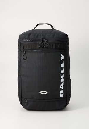 Sort rygsæk med en tekstureret overflade, dobbelt lynlåslukninger, sidelomme og 'OAKLEY' logo i hvidt på forsiden. Kompakt og funktionelt design.
