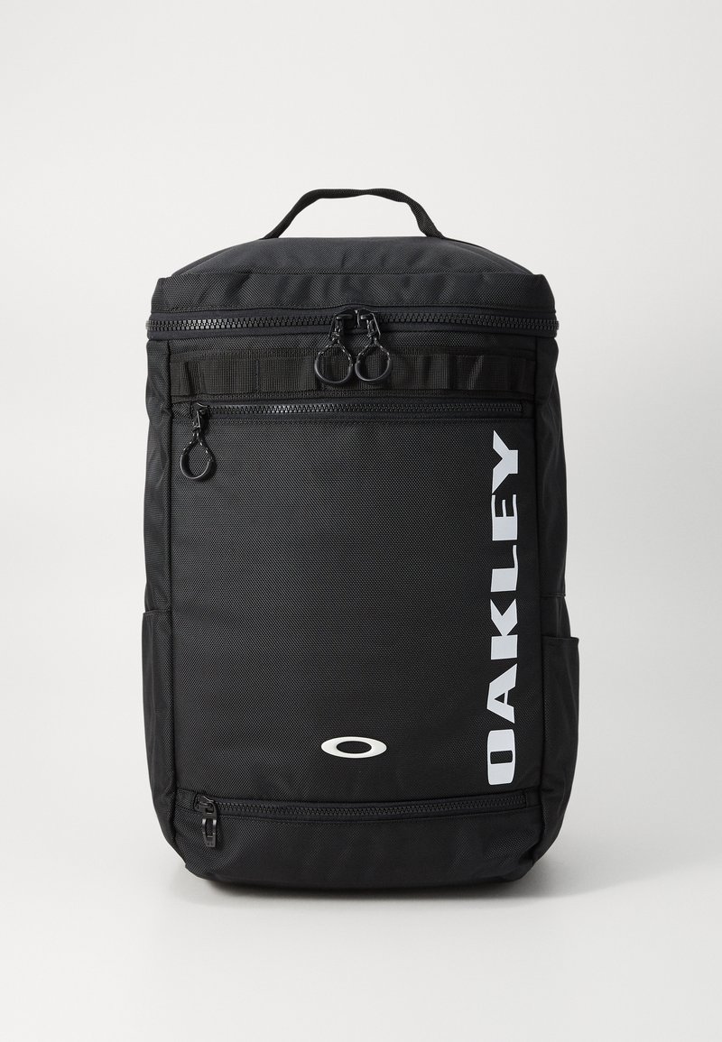 Zwarte rugzak met een gestructureerde oppervlakte, dubbele ritsen, zijvak en een wit 'OAKLEY' logo aan de voorkant. Compact en functioneel ontwerp.