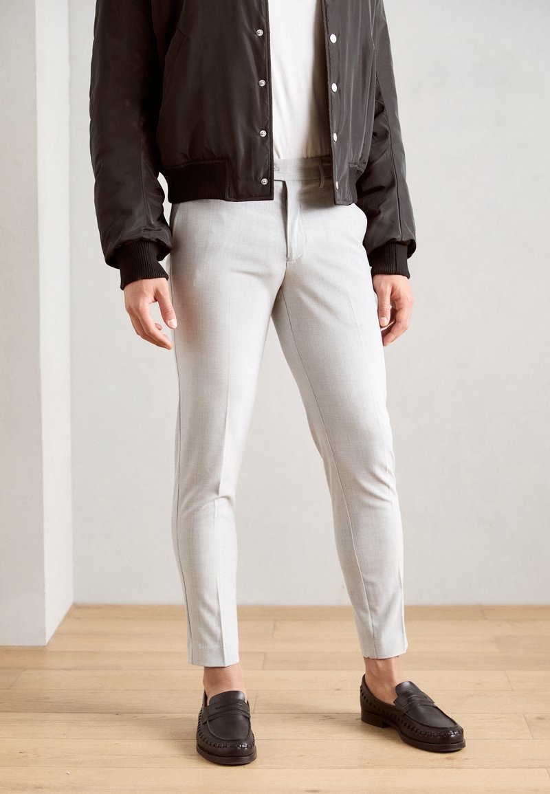 Homme portant un pantalon slim gris clair, des mocassins en cuir noirs, une chemise blanche et une veste noire boutonnée, debout sur un sol en bois.