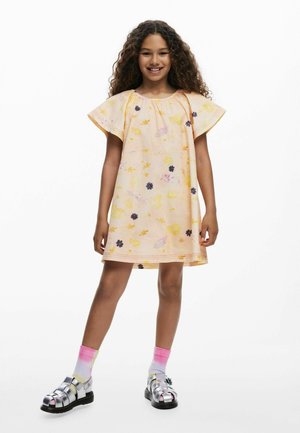 Svjetložuta cvjetna haljina s kratkim, širokim rukavima, ukrašena ljubičastim i ružičastim naglascima. Uparena sa srebrnim sandalama i čarapama u gradijent bojama.