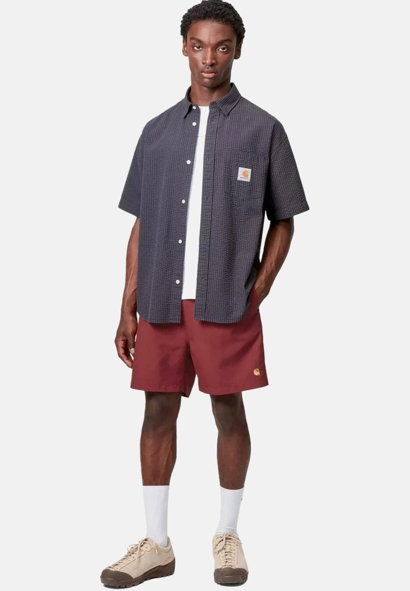 Junger Mann trägt ein dunkel strukturiertes kurzärmeliges Hemd, weißes Unterhemd, dunkelrote Shorts, weiße Socken und beige Sneakers, steht vor einem schlichten Hintergrund.