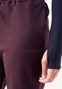Bordeauxfarbene Jogginghosen mit weicher Textur und elastischem Bund, versehen mit dem eingeprägten Markennamen "SWEATY BETTY" am Oberschenkel.