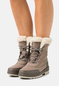 Taupe winterlaarzen met dikke rubberen zolen, crème nepbont voering en grijze veters, gedragen op blote benen tegen een witte achtergrond.
