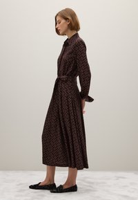 Braun gemustertes Hemdkleid mit langen Ärmeln, tailliertem Gürtel und plissiertem Rock. Schwarze verziehene Ballerinas runden den Look ab.