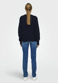 Marineblau gestrickter Pullover mit langen Ärmeln, gerippter Textur, runder Halsausschnitt; kombiniert mit blauen Jeans und weißen Sneakers. Von hinten betrachtet.