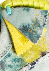 Grønne og gule ruskindspletter med en struktureret finish, der har "CLARKS Originals" branding i guld. Sget detalje er synlig.