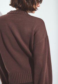 Femme aux cheveux bruns courts portant un pull en tricot texturé marron foncé, vue de dos et de côté sur un fond blanc.