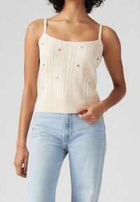 Top de punto crema sin mangas con patrones de trenzado y bordados de estrellas coloridas, con tirantes finos y una cintura ajustada. Combinado con jeans de un azul claro.