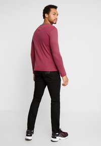 Sarkan un zilu svītrots krekls ar garām piedurknēm un apaļu kakla izgriezumu, apvienots ar melniem biksēm un melniem sporta apaviem. Skatīts no aizmugures.