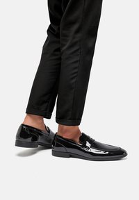 Mocassini in pelle verniciata nera con un design elegante, suola piatta e dettagli cuciti. Abbinati a pantaloni neri arrotolati.