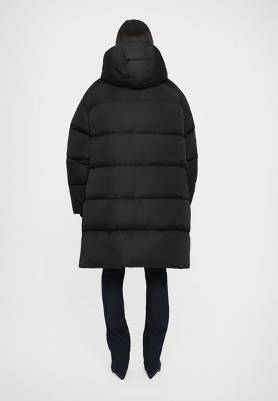 Giacca a vento nera imbottita con un ampio cappuccio e design trapuntato, caratterizzata da strisce orizzontali e una vestibilità comoda, indossata sopra jeans blu scuro.