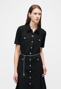 Γυναίκα που φοράει μαύρο φόρεμα με κοντά μανίκια, κουμπιά μπροστά και τσέπες στο στήθος, με ασημένια ζώνη αλυσίδα, στέκεται μπροστά σε απλό ανοιχτό φόντο.