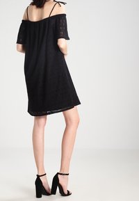 Schwarzes schulterfreies Kleid mit Spitzenstruktur, kurzen Ärmeln und knielangem Saum. Kombiniert mit schwarzen High Heels.