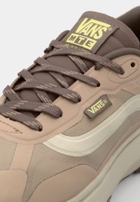Brune Vans sneakers med en teksturert overflate, flate lisser og en hvit side Stripe med en gul logo på tungen. Gummisåle synlig.