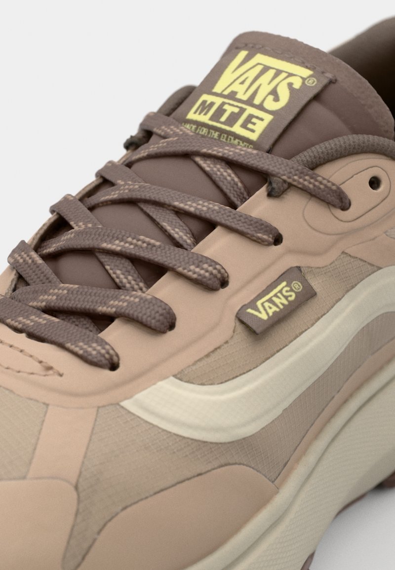 Zapatilla Vans marrón con parte superior texturizada, cordones planos y una franja blanca en el costado con un logo amarillo en la lengüeta. Suela de goma visible.