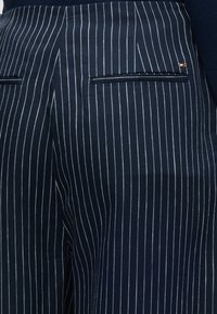 Σκούρο μπλε παντελόνι με λευκές κάθετες ρίγες, διαθέτει δύο πίσω τσέπες με ρέλι, η μία με μικρό ορθογώνιο μεταλλικό λογότυπο.