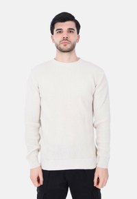 Maglione lavorato a maglia beige chiaro con un motivo a coste testurizzato, scollatura a girocollo e maniche lunghe, abbinato a pantaloni neri.