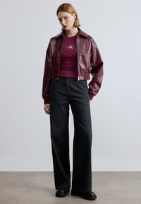 Bordeauxroter Bomberjacke aus Kunstleder mit Kragen, kombiniert mit einem figurbetonten lila Oberteil und weit geschnittenen dunklen Jeans. Schwarze Plateau-Schuhe.