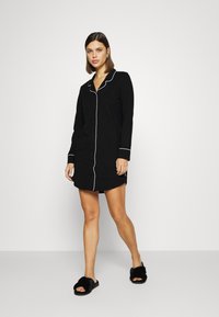 Vestido de pijama preto, com botões, com detalhes em branco, mangas longas e um corte descontraído. Combinado com chinelos fofos pretos para maior conforto.