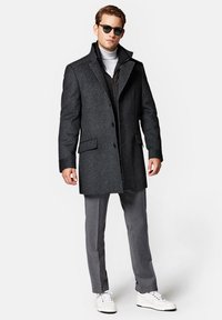 Szary płaszcz z wełny z wysokim kołnierzem, zapinany na jeden guzik, z dwiema przednimi kieszeniami. Nosi się go na czarnej kamizelce, w zestawieniu z szarymi spodniami i białymi trampkami.