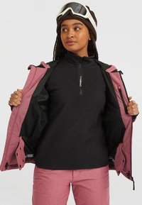Vrouw in een roze skijack en -broek, zwarte fleece trui, bruine gebreide muts en witte skibril, die haar jack openhoudt om de binnenkant te laten zien.