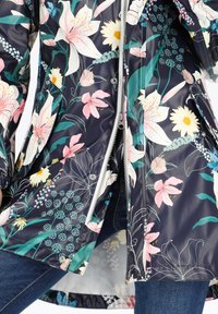 Impermeabile floreale con sfondo blu navy, caratterizzato da grandi gigli rosa e margherite bianche. Realizzato in materiale lucido con chiusura a zip e dettagli a scatto.