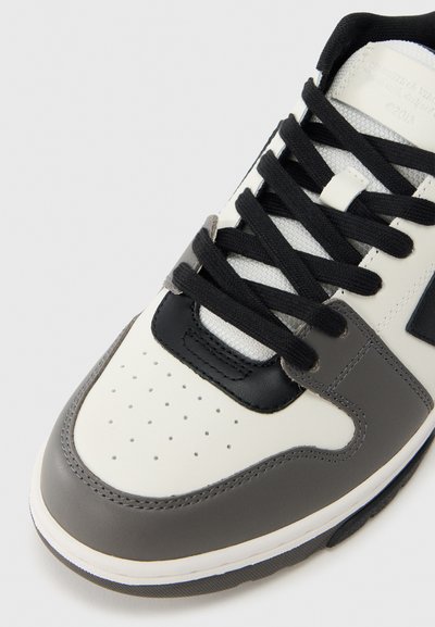 Αθλητικό παπούτσι με λευκό δερμάτινο επάνω μέρος, μαύρες λεπτομέρειες και γκρι επικαλύψεις. Διαθέτει τρύπες στο μπροστινό μέρος και μαύρο δέσιμο με κορδόνια.