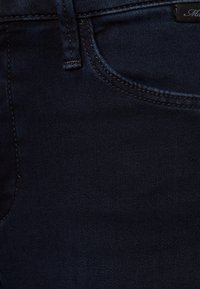 Tissu en denim bleu foncé avec une texture lisse, une légère élasticité et des détails de coutures visibles. Comprend une petite poche avant et une ceinture.