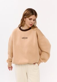 Sweatshirt oversize beige con colletto marrone a coste e logo "MIRONS". Tessuto morbido, spalle scese e orlo rotondo. Abbinato a pantaloni chiari.