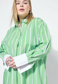 Camicia verde a righe con polsini bianchi, caratterizzata da righe verticali in lavanda e bianco. Realizzata in tessuto leggero con taschino sul petto.