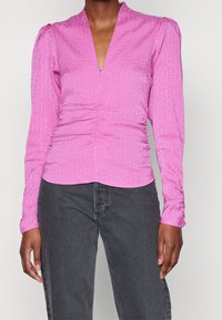 Donna che indossa una camicetta aderente a maniche lunghe di colore rosa con un sottile motivo geometrico e un davanti arricciato, abbinata a jeans grigi scuri.