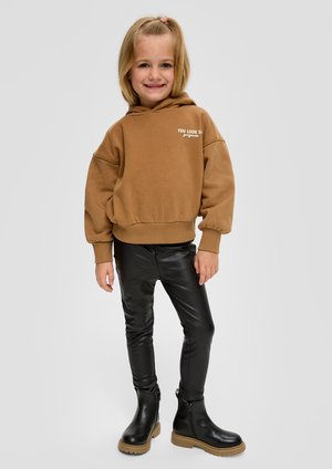 Bruine oversized hoodie met tekst, zwarte nepleer leggings en zwarte enkellaarsjes met een gestructureerde zool. Kort haar met een roze clip.