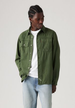 Uomo con capelli intrecciati che indossa una camicia verde abbottonata sopra una maglietta bianca e jeans azzurri, che guarda verso il basso di lato.