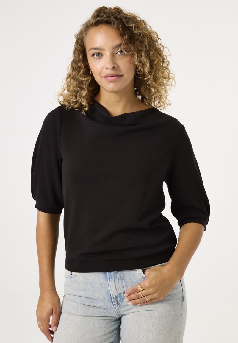 Haut noir à manches courtes avec un col bateau, en tissu texturé et avec une silhouette ajustée. Porté avec un jean taille haute bleu clair.